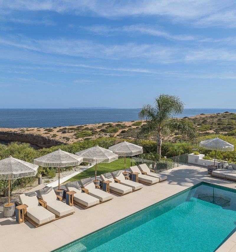 Piscina rettangolare con lettini in legno e ombrelloni con frange affacciata sul mare.